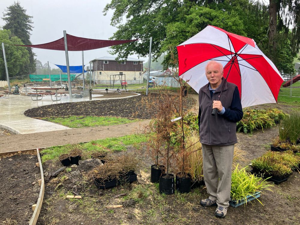 Mr Douglas is a man in his eighties stood next to a beautiful array of native plants with an under construction water play area behind him. He is smiling and holding a red and white umbrella over his head because of course it is raining!