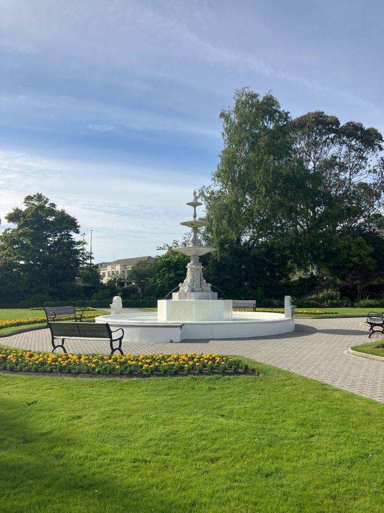 A beautiful green and lush lawn, edged by yellow flowers with a Victorian fountain with multiple tiers happily bubbling away. The sky is a brilliant summer blue. 