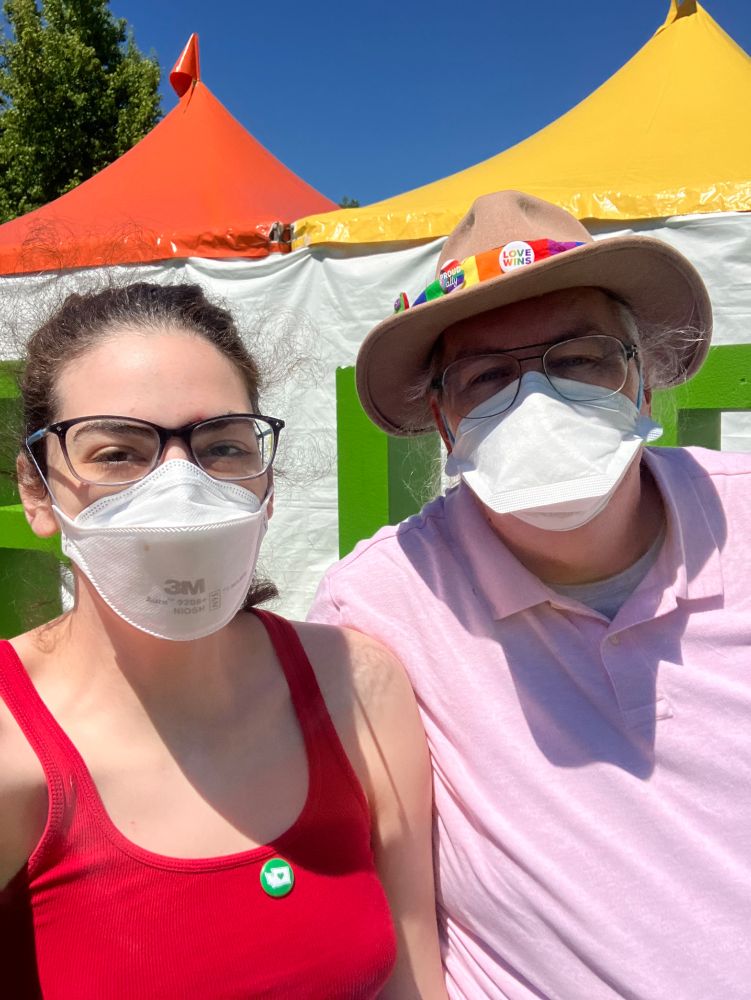 A selfie of Jude and his middle-est daughter at Seattle Pride. Both wear glasses and N95 masks, but their cheeks and eyes show that they are smiling.

Jude is wearing his iconic hat, with a rainbow striped hat band and some button pins on it, and a light pink polo.  

His daughter is a mid-20s white woman, with curly black hair pulled back.  She is wearing a red tank top, with a green button pin that bears a white silhouette of the state of Washington.

In the background, there are obstructed tall green letters on a white background, and two tent roofs from the Seattle Pride sign, one yellow, one orange.