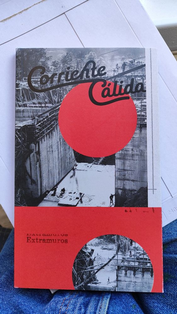 Fotografia d'una revista gruixuda sobre un paquet de missatgeria sobre la falda.
A la revista hi ha una fotografia en blanc i negre amb formes geomètriques vermelles. 
A la revista hi posa: 
Corriente Cálida
Extramuros
