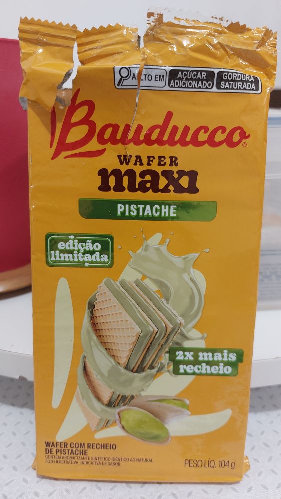 Embalagem amarela do wafer Bauducco Maxi Pistache com destaque para um wafer branco com um recheio verde claro. Lê-se "ex mais recheio" e "edição limitada"