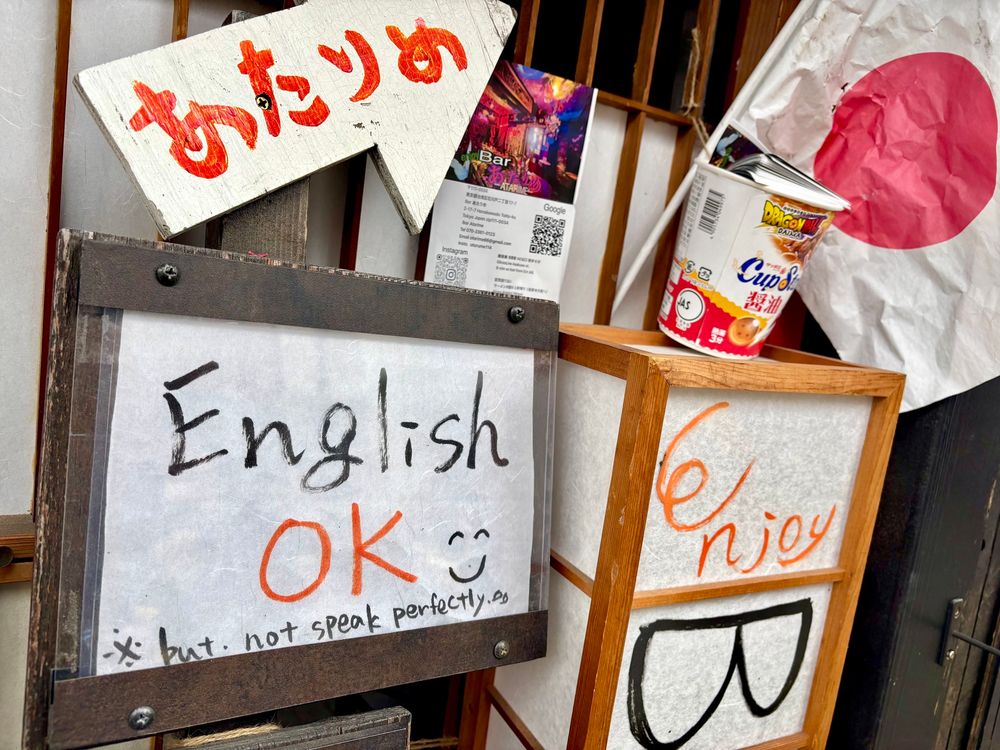 Outside a small bar in a tourist district. A Japanese flag, a menu, a shoji lantern, a cup noodle (I think), and a sign that says English Ok with a smiley face but with fine print on the bottom that says *but not spoken perfectly. 