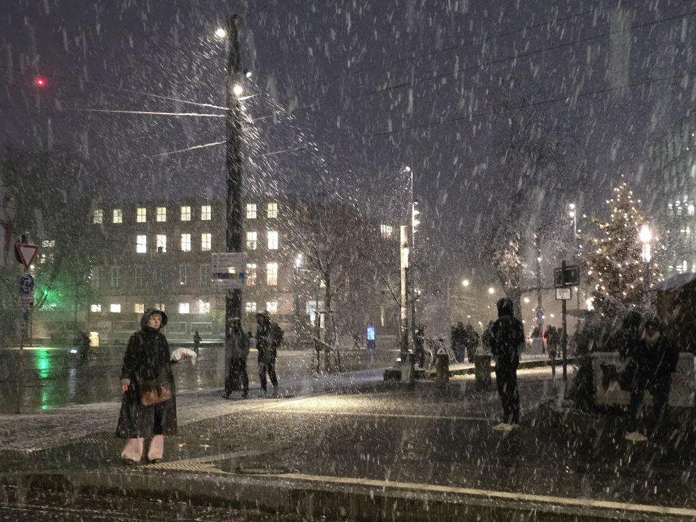 Light snow on the streets of Freiburg. Some figures are seen walking or standing on the sidewalks. 