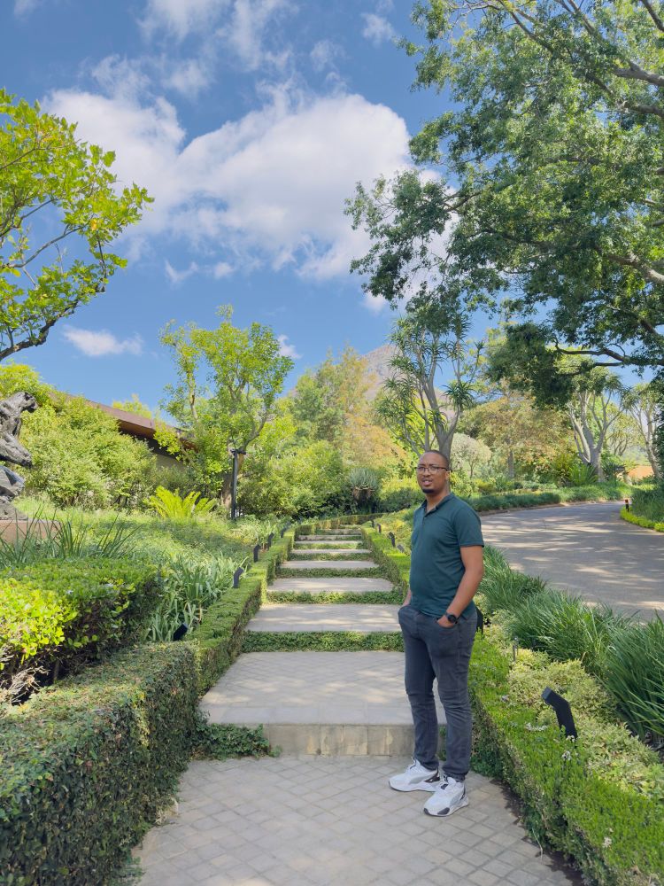 Phila Msimang standing with his hands in his pockets on some stairs surrounded by green trees and shrubs. 