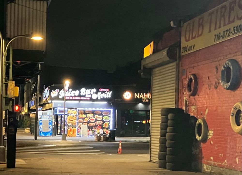 A nighttime shot of a Brooklyn intersection. In the foreground is a tire shop with some tires stacked up beside the closed garage. Beyond is the Ave P Juice Bar and Grill, which was still open and makes excellent fresh sandwiches 
