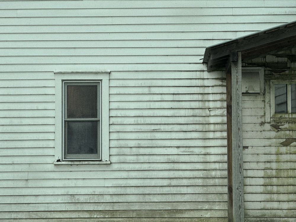 This photograph shows the side of a white house with horizontal siding, weathered with streaks of dirt and mold. On the left side is a closed, double-hung window with a dark mesh or screen, while the right side reveals a corner of a porch roof supported by a single wooden post. Faint green and brown discoloration marks the siding near the porch, where a second, smaller window and some peeling paint suggest long exposure to the elements.