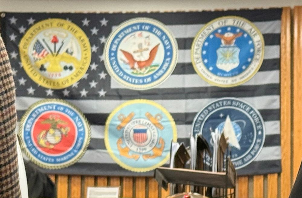A very large black and white American flag with the logos for the Army, Navy, Air Force, Marines, Coast Guard and Space Force imposed on top of it. 