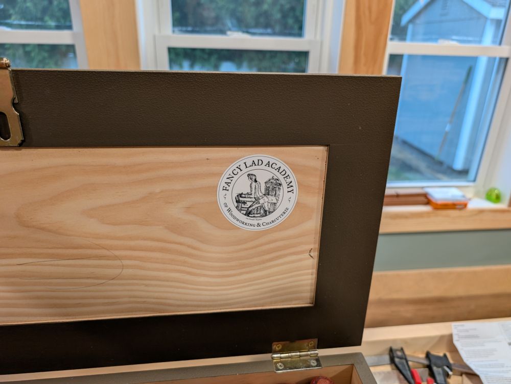 The inside of a small wooden tool chest and blazoned with a sticker that reads "fancy lad academy of woodworking and charcuterie" originally created by Lost Art press. 
