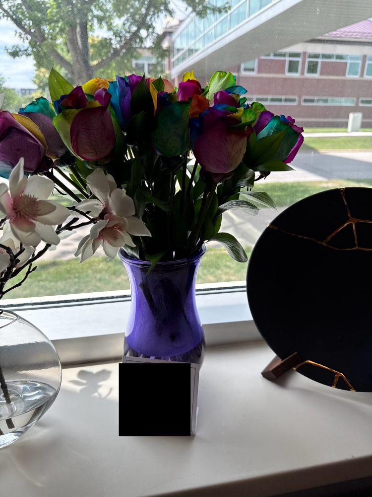 A bunch of rainbow colored roses in a blue vase next to a decorative Japanese kintsurigi plate on an office window sill. 