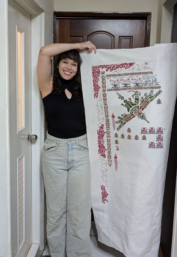 A person standing and holding up a large cloth with parts of a tapestry stitched on kt