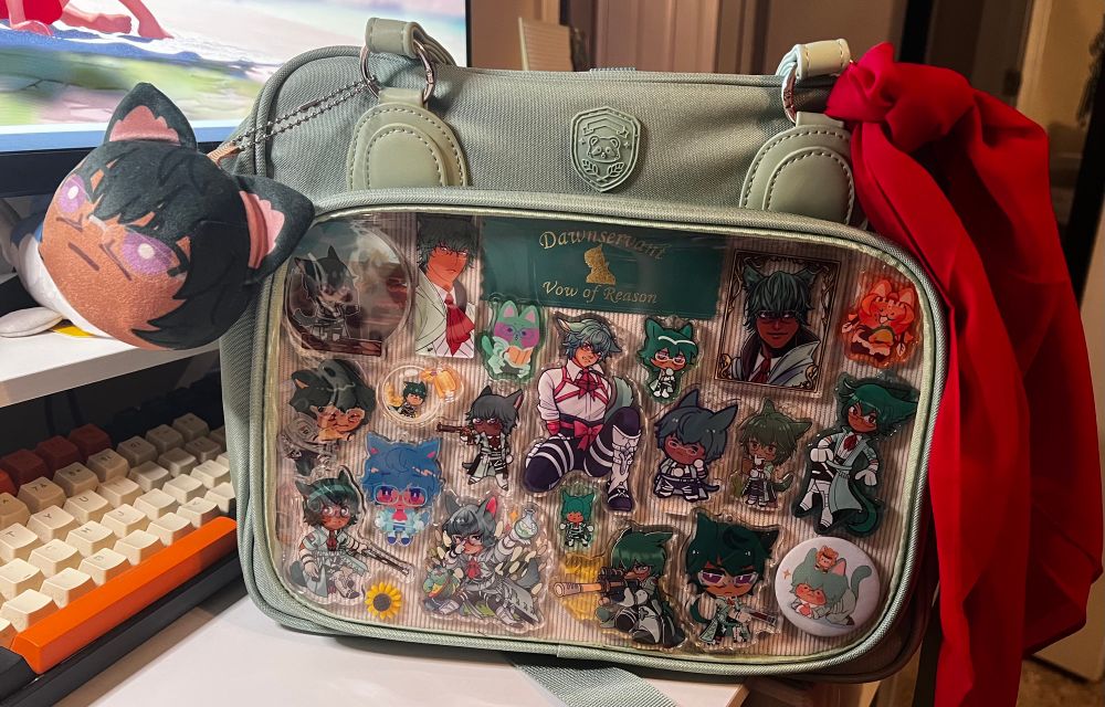 A photo of a light green itabag with a front window displaying various charms of koana from final fantasy xiv. There is a Koana omanjuu hanging off one strap and a red neck tie similar to the one Koana wears tied to the other. 