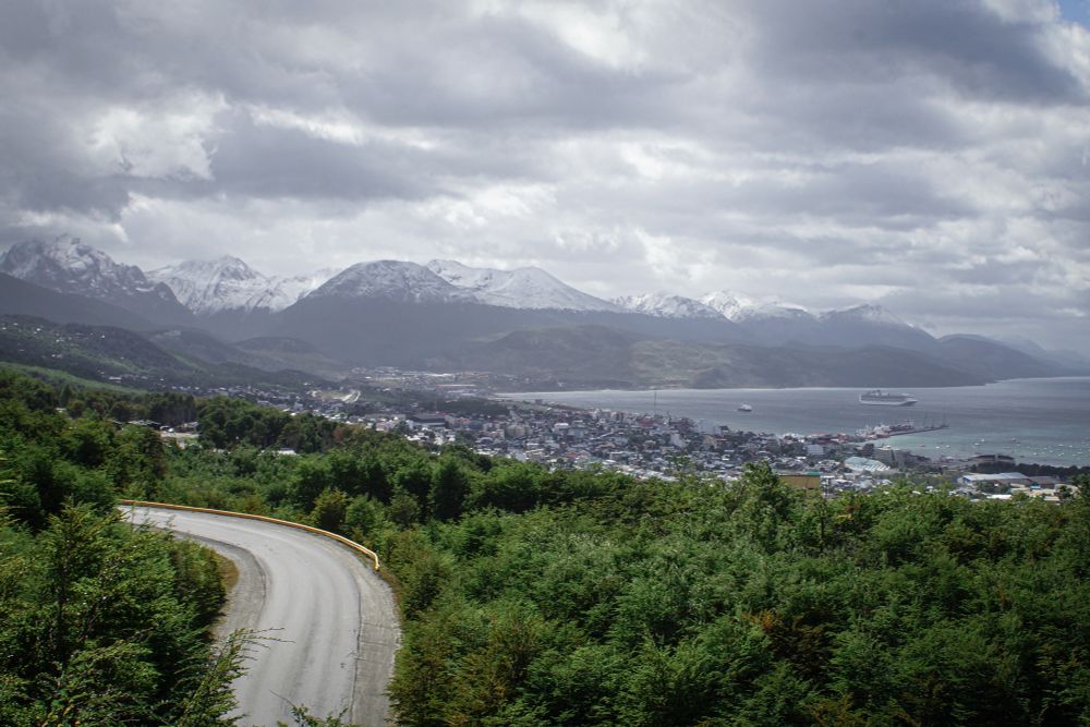 Panoramic view of Ushuaia, Argentina, with mountains in the background and greenery in the foreground. Ushuaia is nestled in the middle by the bay.