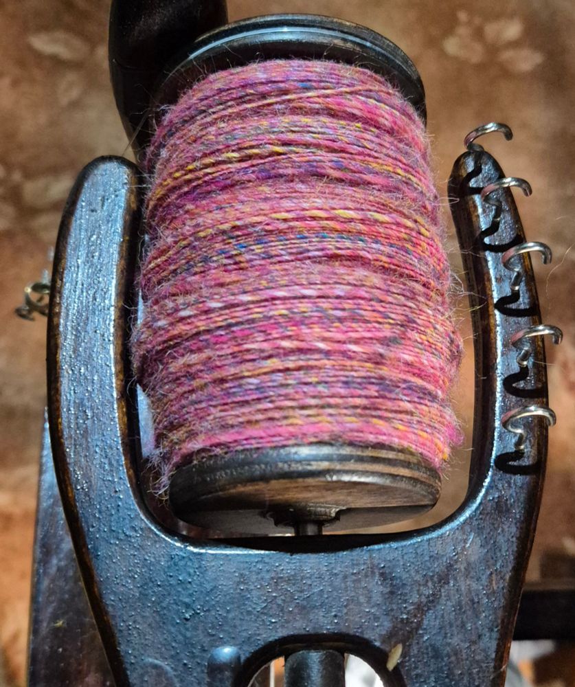 A bobbin full of mainly pink handspun fibre shown still on the spinning wheel flyer. The pink spun fibre has blue, yellow and white shot though it. The background behind the wheel is an orange carpet.