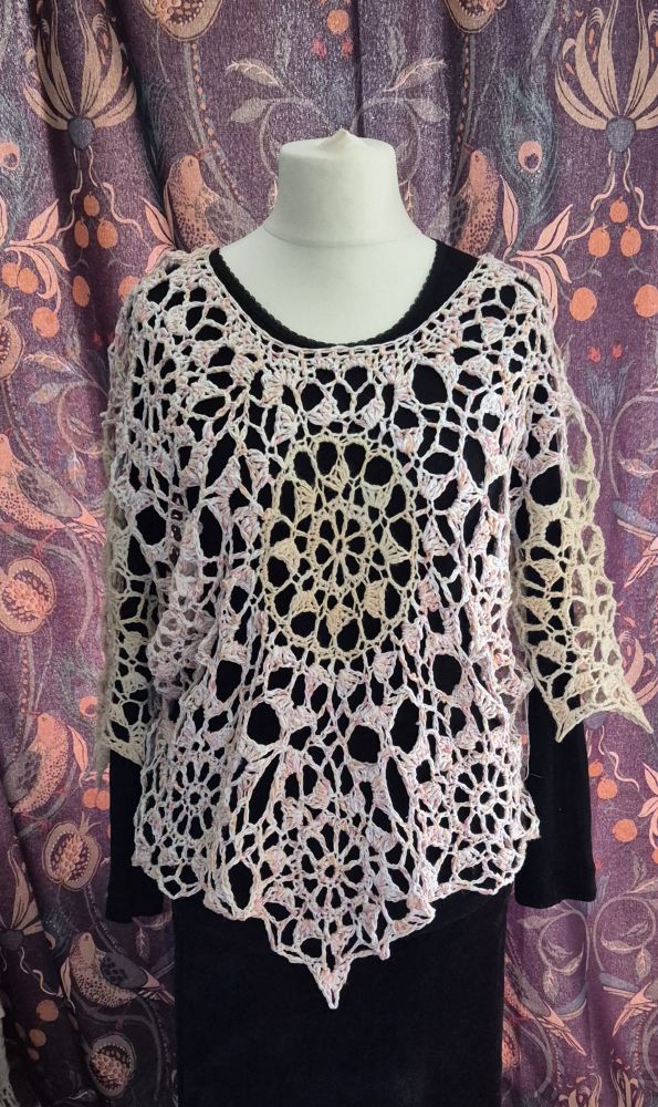 The front view of a handmade pink and cream coloured crocheted top layered over a black top and skirt set against  a blue and colourful curtain.