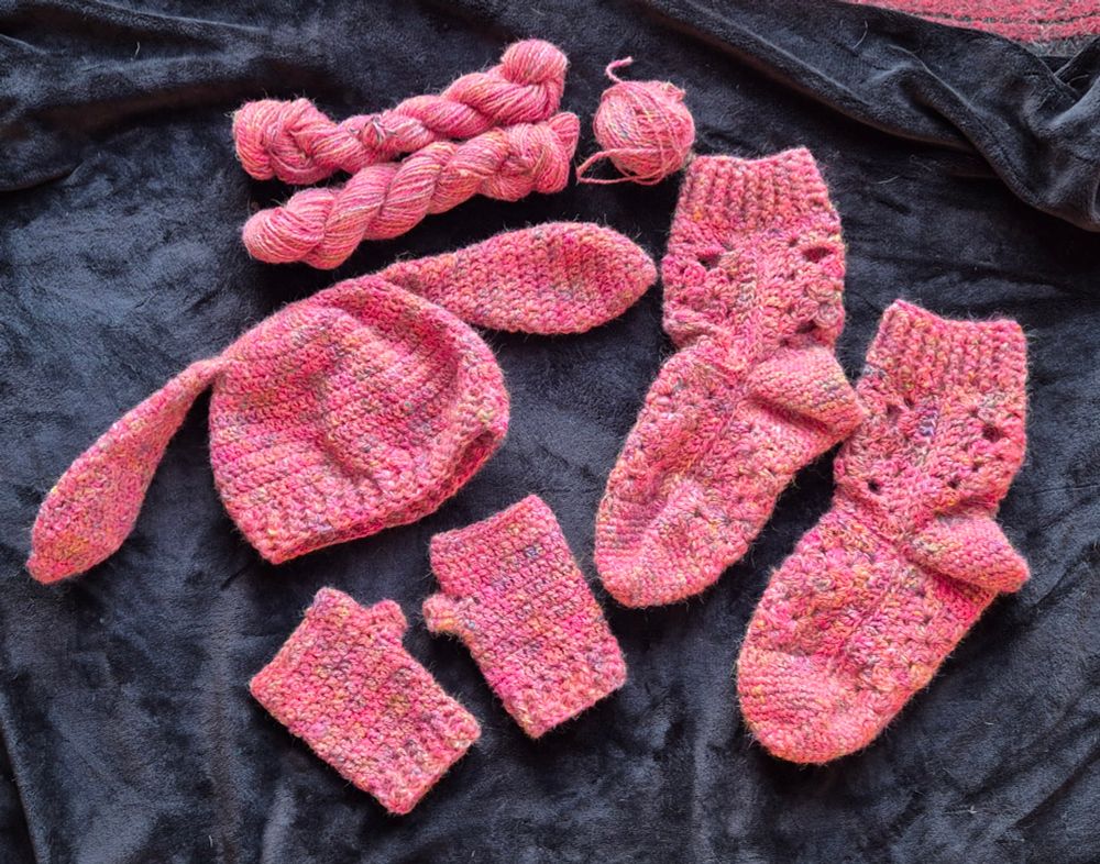 Picture shows a pair of  crocheted socks, a pair of crocheted hand mittens, a crocheted hat with bunny ears and a ball of two skeins of plyed wool. All of the wool and crocheted projects are made with pink handspun wool. All set against a black background 