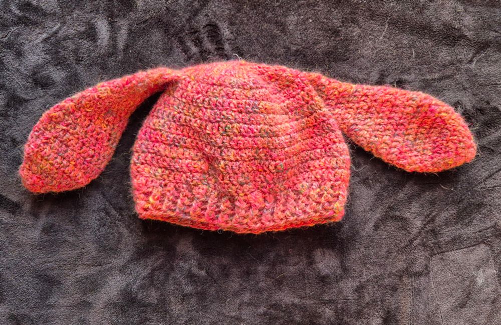 Crocheted hat with ears using pink handspun yarn set against a black background 