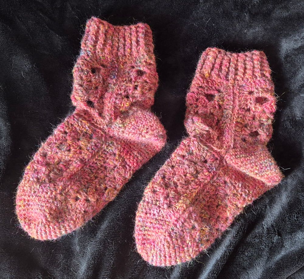 A pair of granny square crocheted socks. The socks are made with a pink handspun wool. The socks are set against a black background 