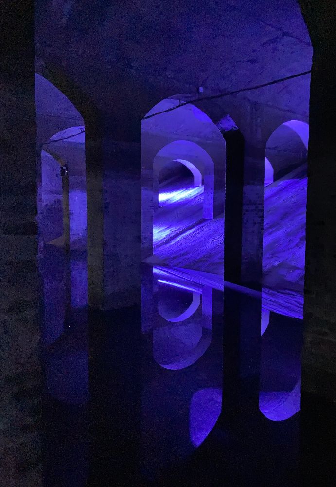 A blue light installation illuminating arched vaults in an underground space. The floor is covered in perfectly still water that reflects the vaults and lights. 