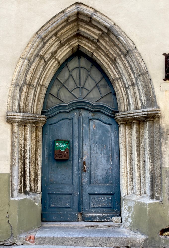 A photo of a blue wooden double door in a medieval gothic arched doorway. There’s a decorated window above the door to fill out the pointy upper part of the doorway. A planter with something green in it is hanging on one half of the double door, likely the half that doesn’t get opened. 