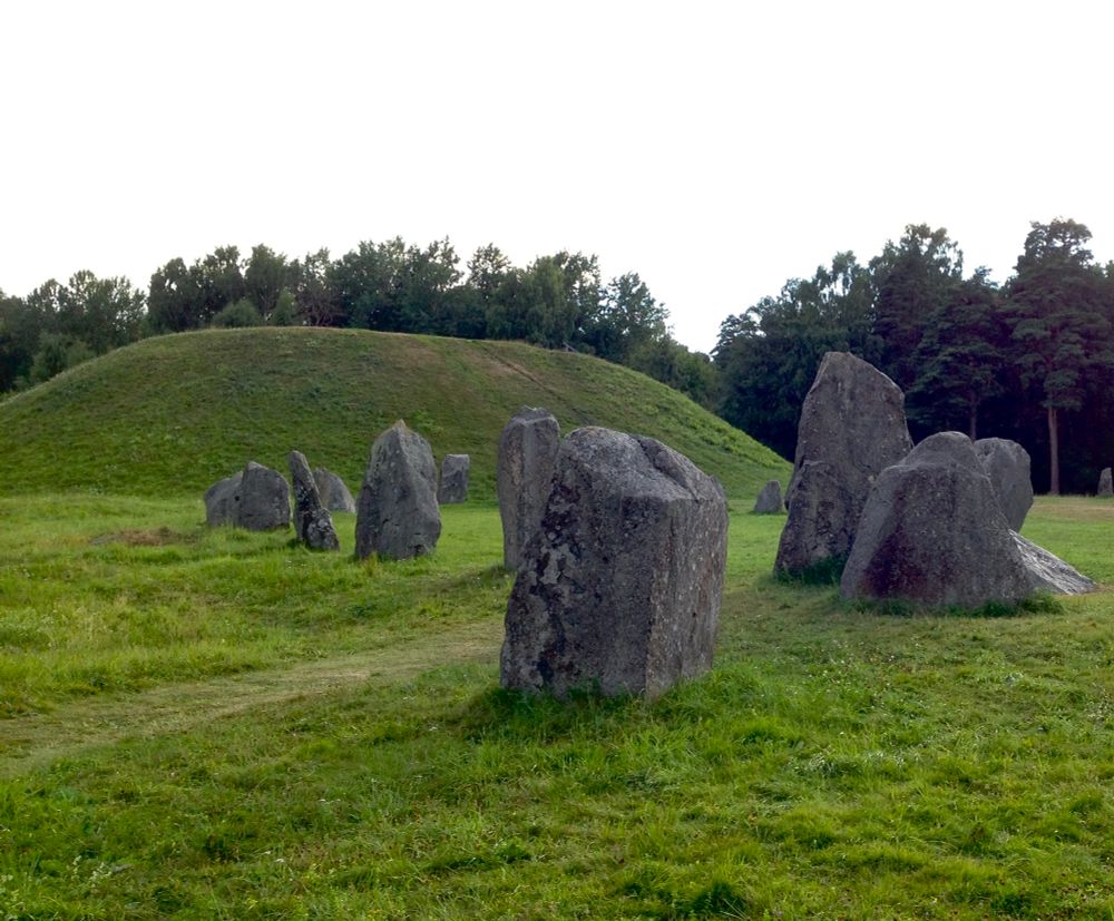 A photo of a grass covered mound wit some standing irregular grey boulders in the foreground. 