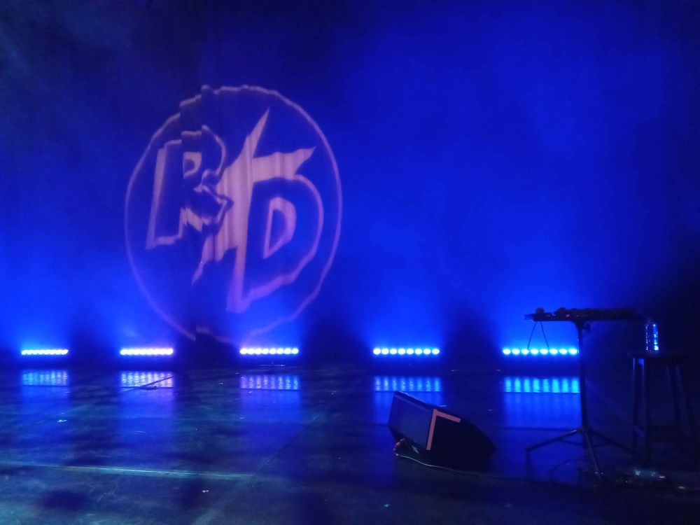 Photograph of the stage of Rhys Darby's show at the Edinburgh Fringe, with a beatbox doohickey on a table and his logo projected on the curtain backdrop