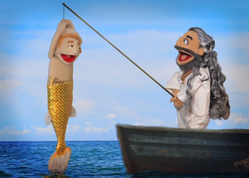 A photograph of two muppets at sea. The Ed Teach muppet is in his rice sack onesie and he's holding a fishing line. He has caught a merStede muppet who is dangling from his fishing line and they are both staring at each other in delight.