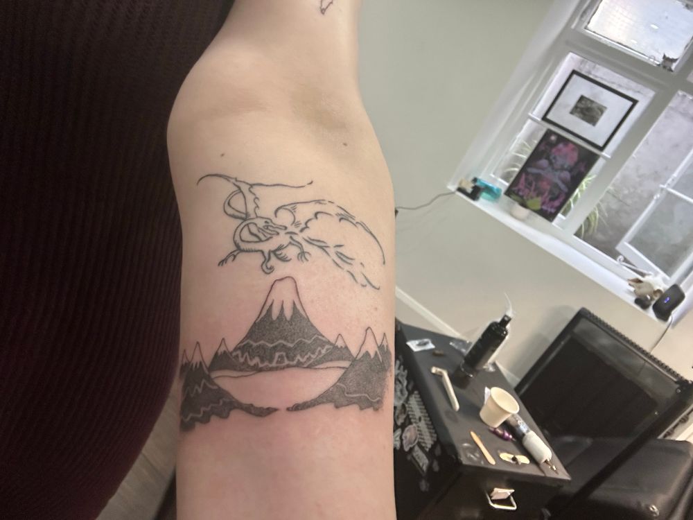 Tattoo of a dragon breathing flame flying over a range of mountains from the cover of Tolkien’s The Hobbit