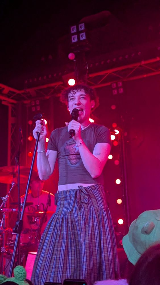 Nemo on stage wearing a tartan skirt and a crop top with heart on it, smiling while they sing