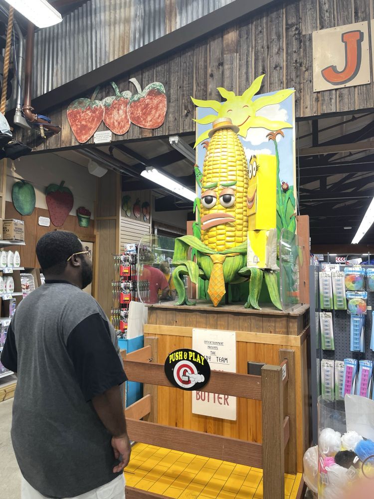 A photo of Benny observing an animatronic corn and stick of butter with faces at Jungle’s Jim. Also the corn is wearing a tie.