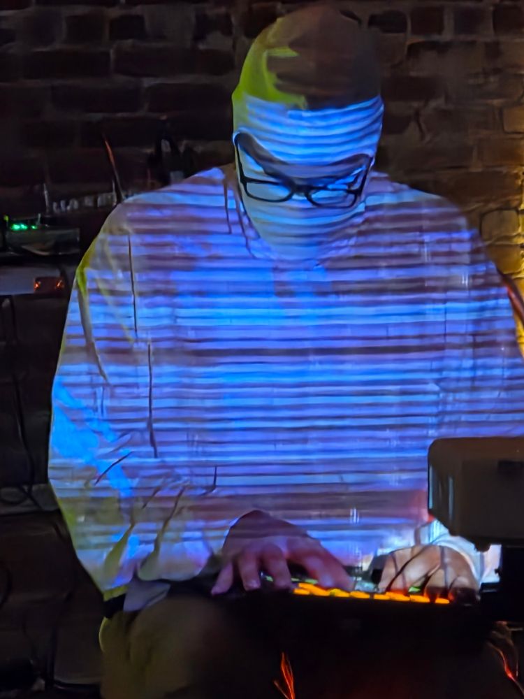 Photo of a guy with video of blue scan lines projected onto his torso and face, mid-performance, looking down, hands on a lighted midi interface.
