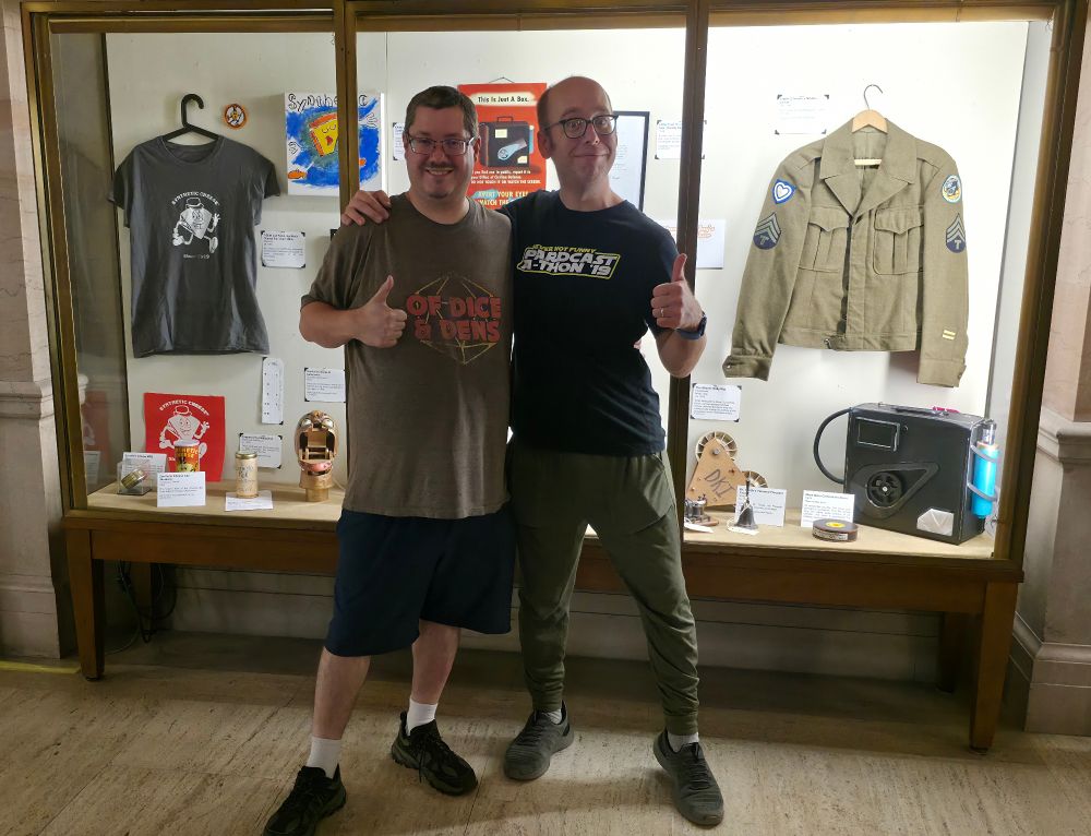 Two nerds in walkin' 'round attire, thumbs-upping the camera with a museum style display in the background 