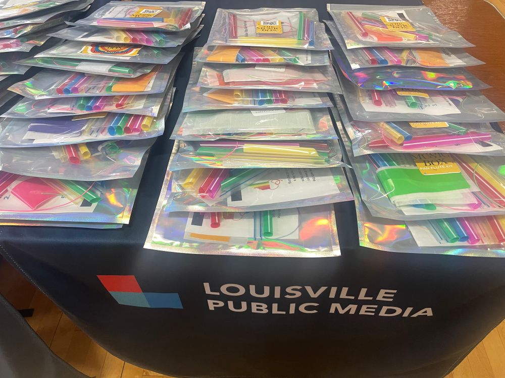 A table is laid out with flute-making kits, featuring multicolored straws and tape. A tablecloth beneath the kits shows the logo for Louisville Public Media.