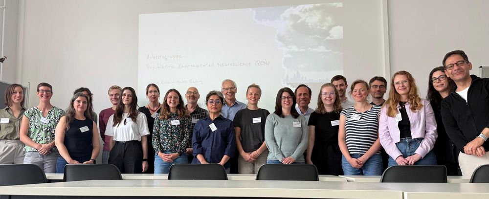 A group of 21 researchers of different ages and gender stand in a row behind a long table in a conference room. They are all facing the camera and smiling, many with their arms held in front of them. Behind them is a screen with the words "Arbeitsgruppe Psychiatric Environmental Neuroscience (PEN) projected onto it. 