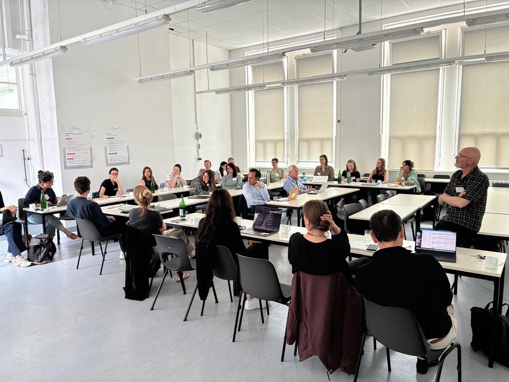 A group of researchers are gathered in a white-walled conference room or classroom. The researchers, mostly clad in dark casual clothing, sit at right-angled tables, and turned to face someone in the back row, who appears to be speaking. They look serious and in concentration. 