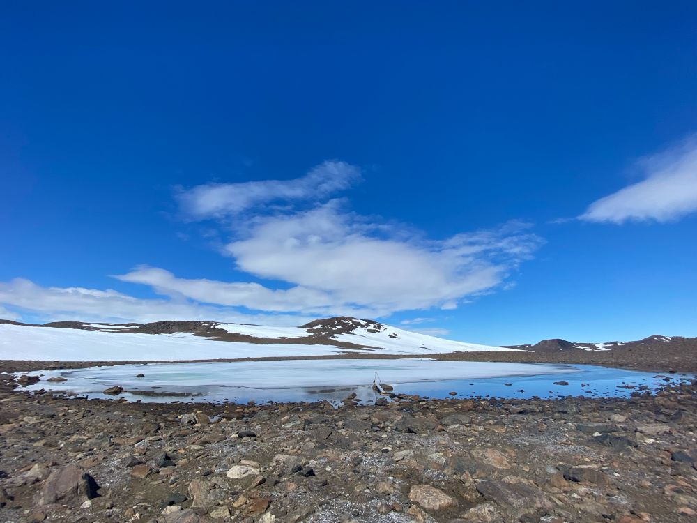 A semi frozen lake is surrounded by a rocky shoreline. In the distance are snow covered hills. The sky is bright blue with some fluffy clouds. This is our water supply 