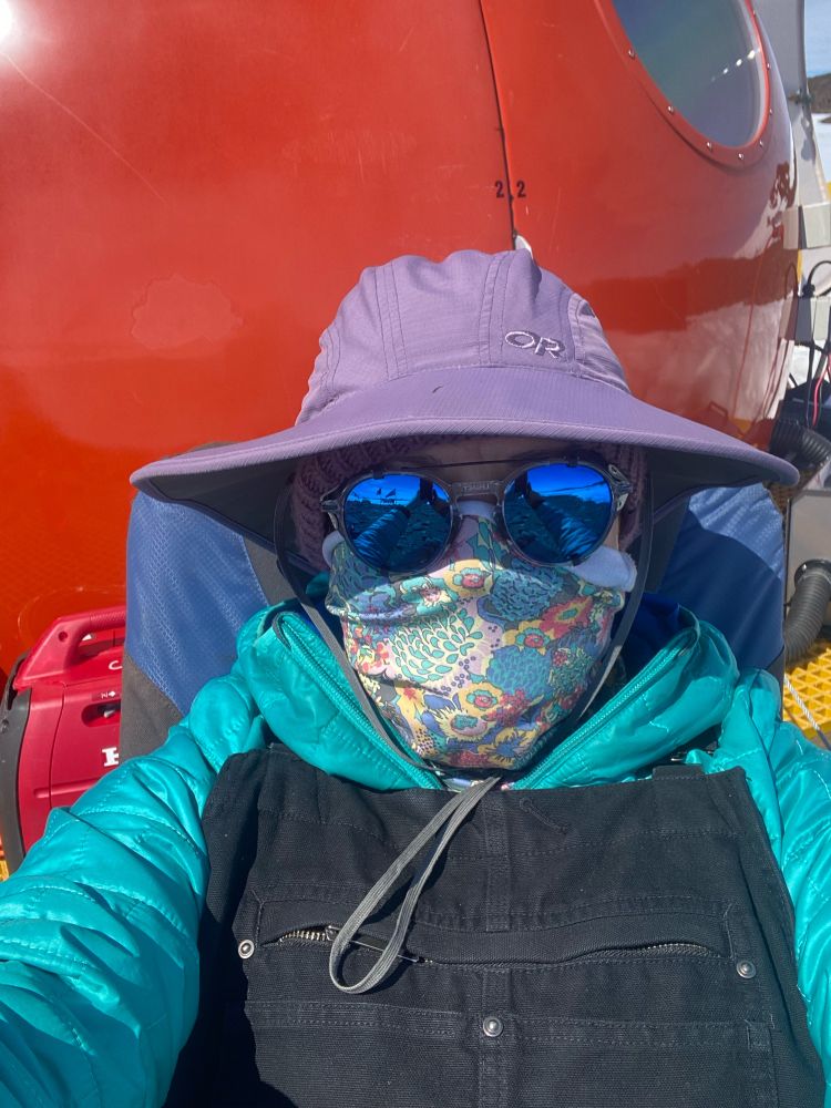 A selfie of a woman with a floral face covering, blue glacier glasses, purple wide brim sun hat, teal microdown jacket and black carhart overalls sitting in a deck chair in front of a red fibre glass melon building 
