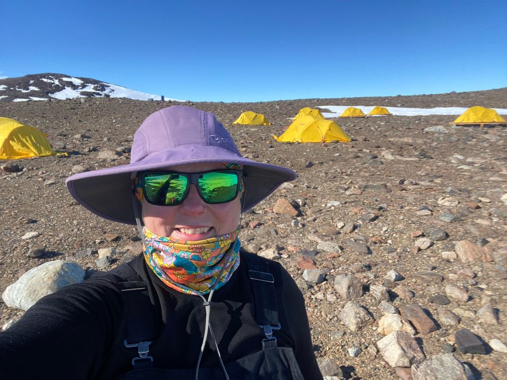 A white woman jn a purple hat and sunglasses and black polar clothjng stabds in front of the rock hills of Bunger Hills. In the background are 6 bright yellow polar dome tents. This is the Edgeworth-David field camp in the bunger hills 