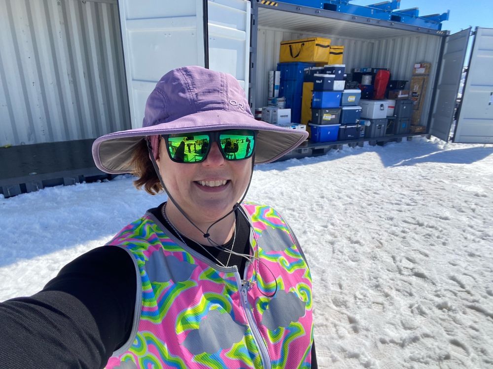 A white woman wearing a purple sun hat, green reflective glasses and a fluorescent rainbow cloud high vis vest over a black thermal top stands in front of a side opening shipping container filled with hard plastic cases for science equipment. It’s a bright sunny day, and there’s snow on the ground with pours of foot prints in it 
