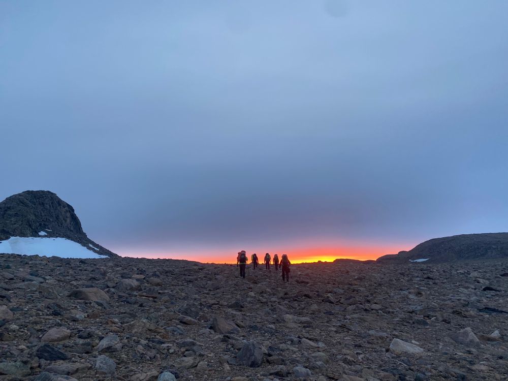 The silhouette of 5 figures walking up a rocky slope towards a glowing band of sunrise. Grey clouds in the sky, to the left is Camel’s Hump (a rock formation) at Bunger Hills, Antarctica 