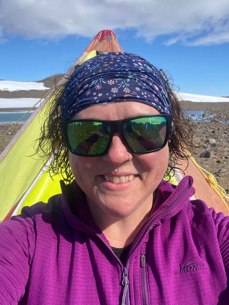 A white woman with short brown curly hair and a purple jumper and sunglasses stands in front of a polar pyramid tent. Her hair is freshly washed 