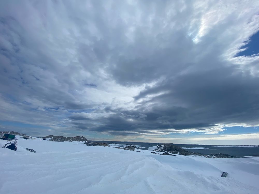 Dramatic skies over Casey! There’s grey storm clouds with some sunshine out at sea. In the foreground is sastrugi snow rolling down to dark rocks and the navy blue ocean 