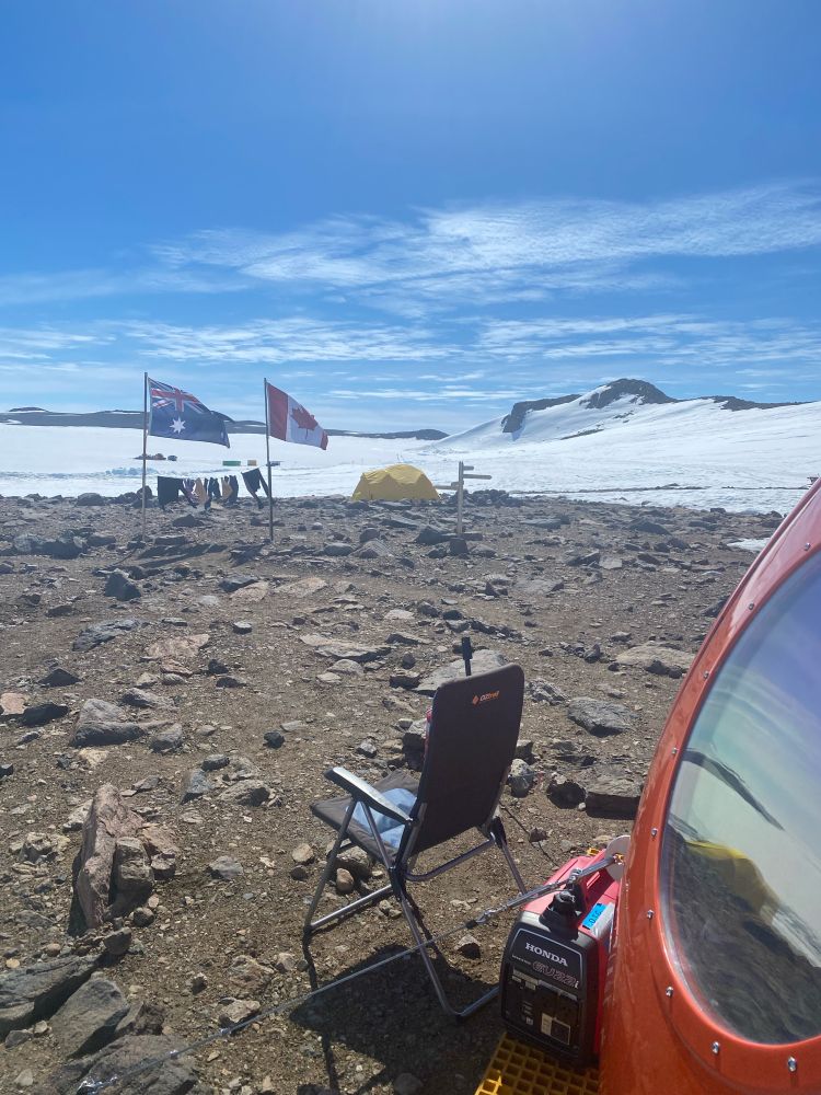 The circular window of the red fibreglass melon hit, a blue deck chair on rocky ground overlooking an Australian and Canadian flag with washing stung up between the poles. In the background a yellow polar dome tent, and the sea ice 
