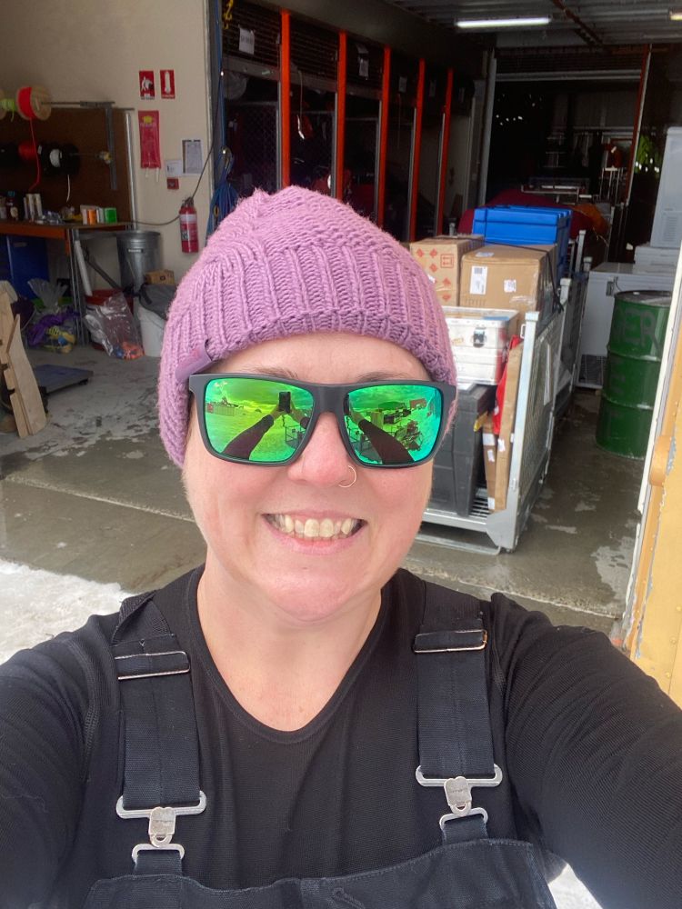 A selfie of a white woman wearing a purple cable knit beanie, green reflective glasses, and black overalls and thermal top. She stands in front of a yellow building with cage pallets of equipment in the open door 