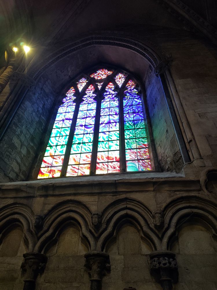 The Illumination Window: a large, rainbow coloured stained glass window in Durham cathedral dedicated to the memory of student, Sara Pilkington. It has a modern pattern with hundreds of abstract shapes that look like birds rising from the land and sea