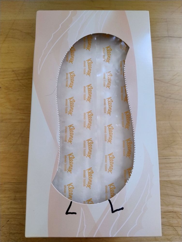 A Kleenex tissue box photographed from above, opened by tearing away the perforated section of the box top. The cut-out section is shaped like the body of a wug. Two wug legs have been drawn on the box, at the bottom of the cut-out section.