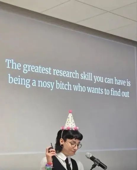 A thin white person with a dark mullet hairstyle, glasses, and a party hat stands in front of a PowerPoint projection that reads: "The greatest research skill you can have is being a nosy bitch who wants to find out".