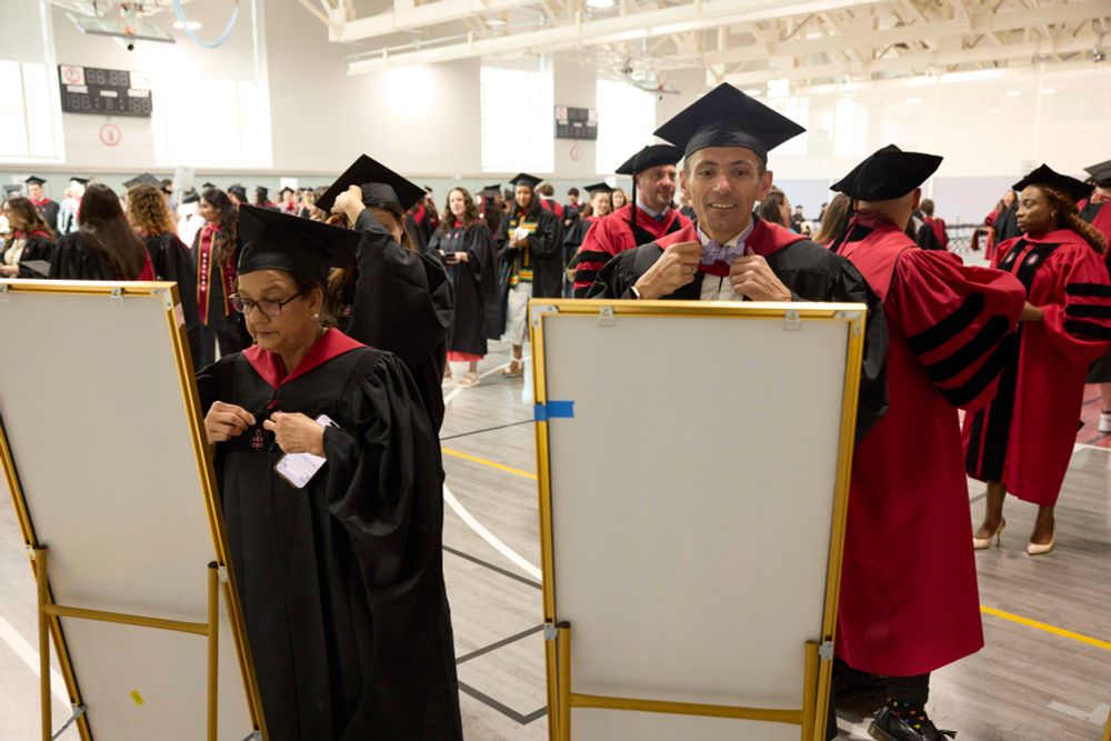 Graduates in red and black academic regalia prepare for the Harvard Chan School 2025 Convocation, adjusting their outfits in front of mirrors.