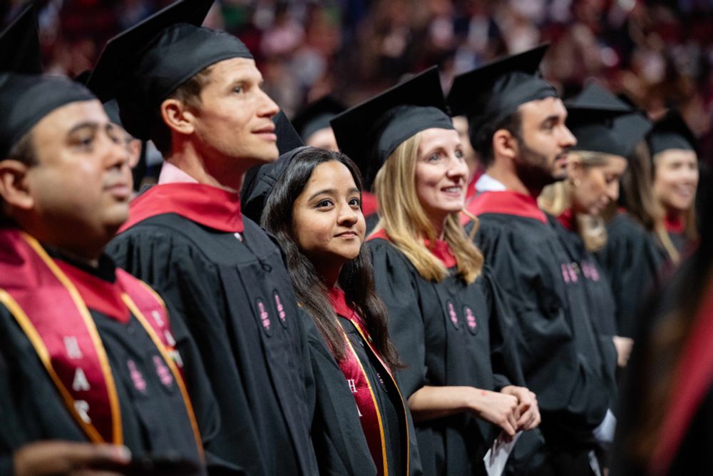 Graduates wearing caps and gowns at the Harvard Chan School Convocation ceremony, looking attentively in the same direction with smiles.
