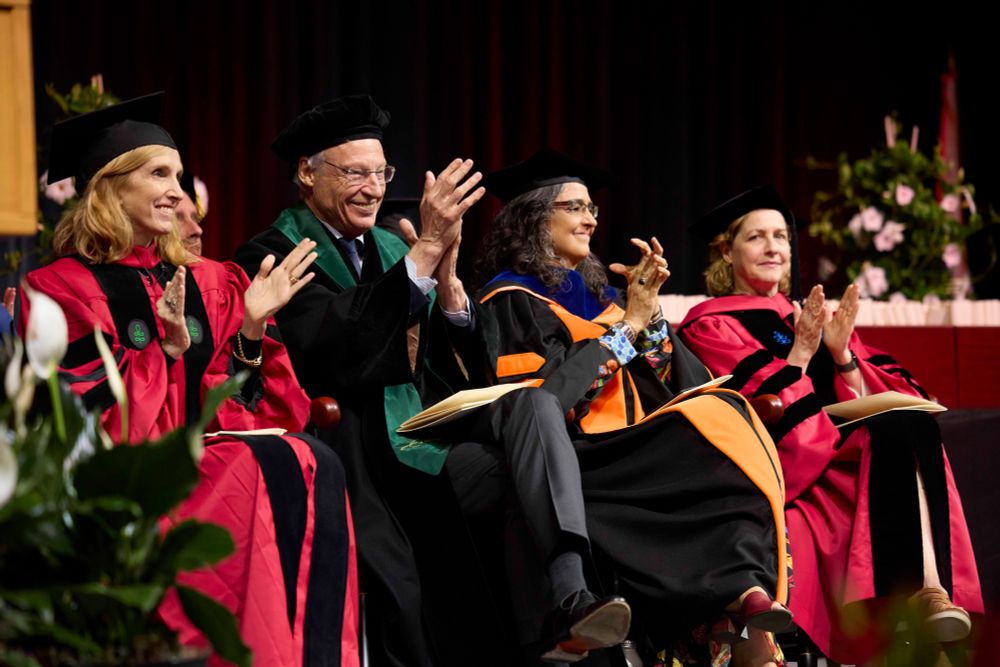 Harvard Chan faculty members in traditional regalia, seated onstage during the Convocation ceremony, clapping and smiling.