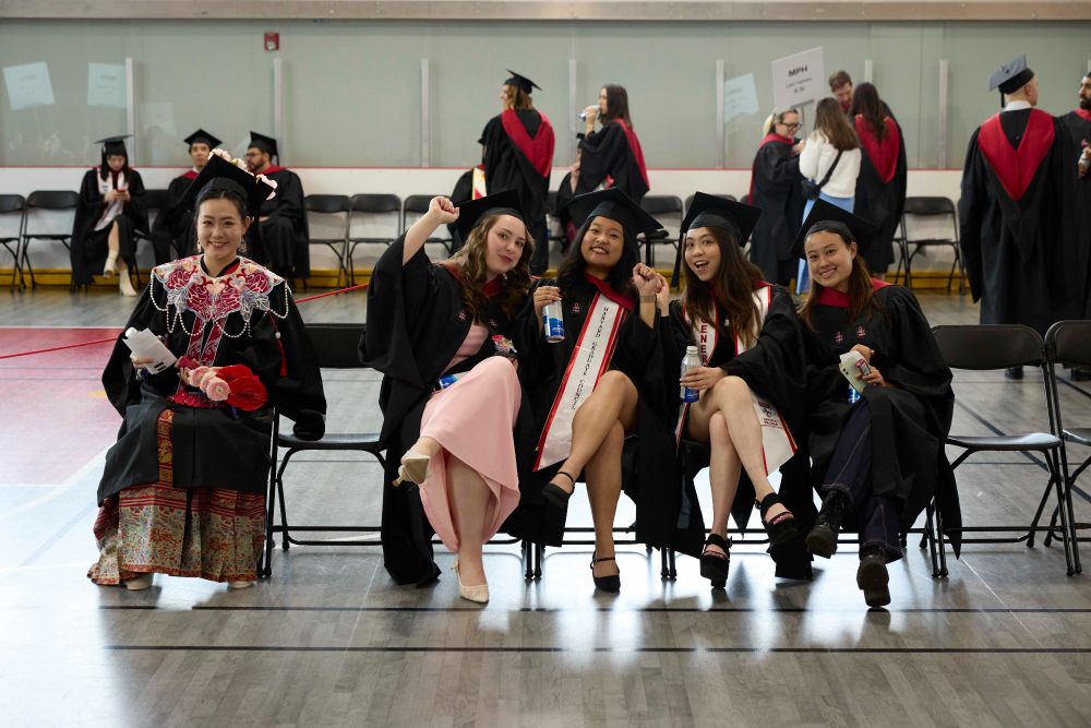 A group of graduates in caps and gowns, sitting together and celebrating, each displaying a joyful expression. Some are waving at the camera and holding diplomas.